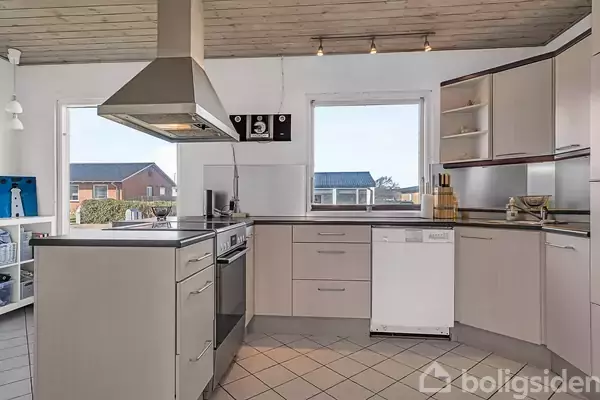 Køkken med kogeø og emhætte, omgivet af lyse skabe og skuffer. Store vinduer tillader naturligt lys, med udsigt til nabohuse og have. Flisegulv og træloft fuldender rummet.