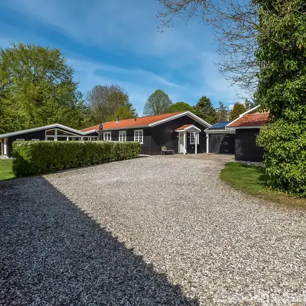 Et sort hus med rødt tag står i en grøn have med grusopkørsel. Træer omkranser huset, der ligger i et fredfyldt boligområde under en blå himmel.