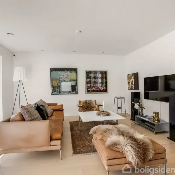 En lys stue med lædersofaer og en chaiselong dækket med et pelsbetræk. Et fladskærms-tv er monteret på væggen, omgivet af kunstværker. Store vinduer giver udsigt til en terrasse.