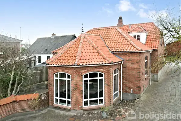 Rødt murstenshus med buede vinduer og rødt tegltag står langs en brolagt sti. Omgivet af andre huse og et hegn i en villakvarter.