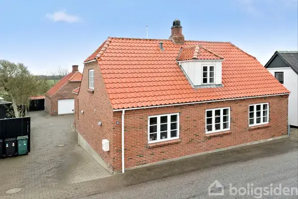Murstenshus med rødt tegltag står på en brolagt gårdsplads. To etager med kvist og skorsten. Omgivet af landlig baggrund med forårsskov og blå himmel.