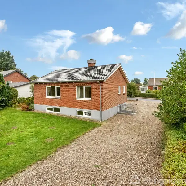 Et rødt, murstenshus står på en grusvej i en frodig have med træer og græsplæne. Andre huse er synlige i baggrunden under en blå himmel med skyer.