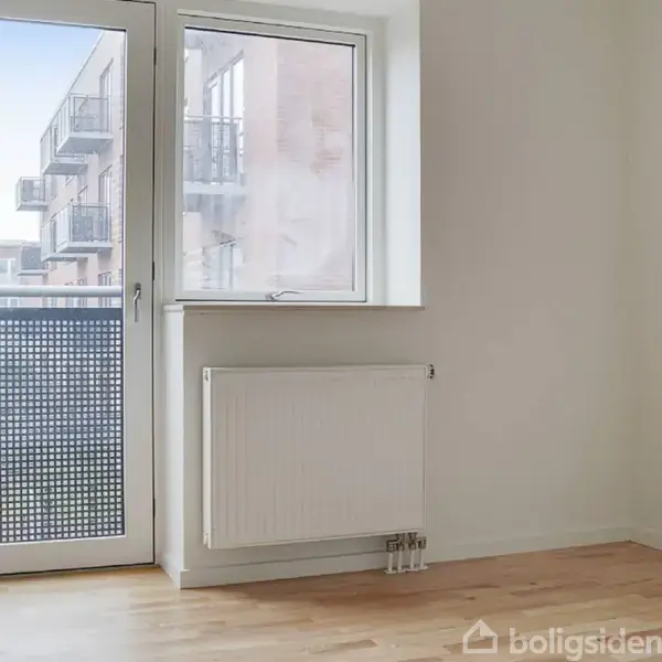 En lys trædør og et vindue med radiator under, åbner til en altan. Udendørs ses en moderne bygning med flere altaner. Indendørs er der trægulv og hvide vægge.