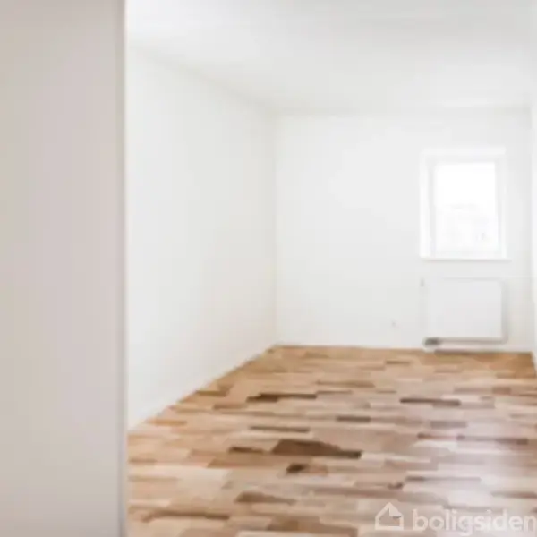 Et tomt værelse med lyse trægulve og hvide vægge har et vindue og en radiator på den fjerneste væg, hvilket giver et minimalistisk og lyst udtryk.