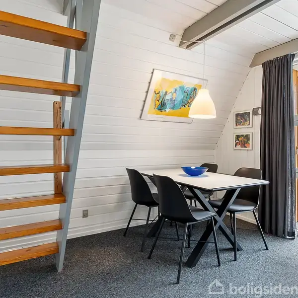 En trappe af træ og metal står ved siden af et spisebord med fire stole i en lys stue. Store vinduer viser en terrasse og grøn natur udenfor.