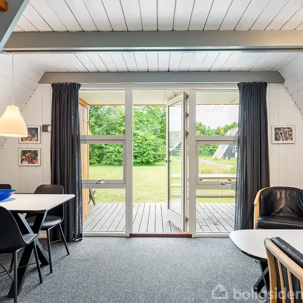 En stue med spisebord og stole står ved siden af en lænestol. Glasdøre åbner mod en terrasse og grøn have. Væggene er træhvide, og der hænger en lampe over bordet.