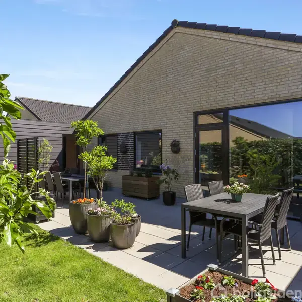Et moderne hus med fladt tag og lyse mursten står i en have. En terrasse med plantekrukker og sorte havemøbler befinder sig foran huset.