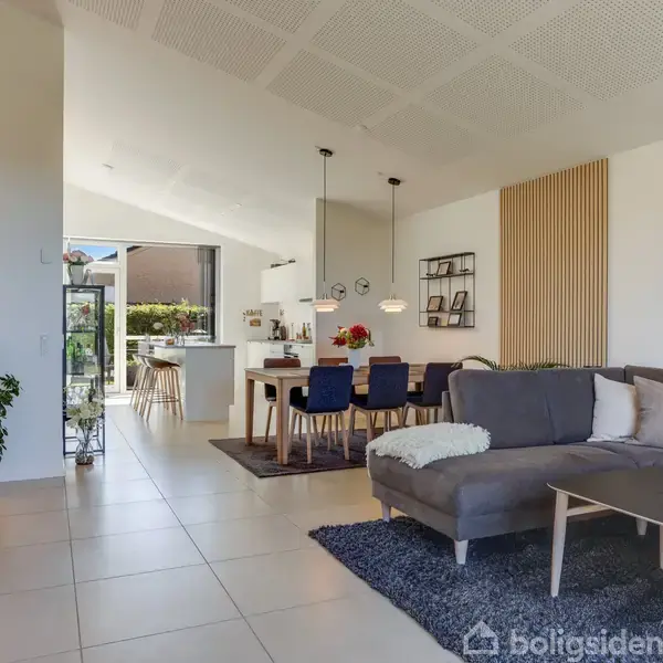 Stue med grå sofa og sofabord, pejs på venstre side. Spisebord med stole i midten, hængende lamper over. Baggrund med køkken, lyst og moderne interiør, planter omkring.