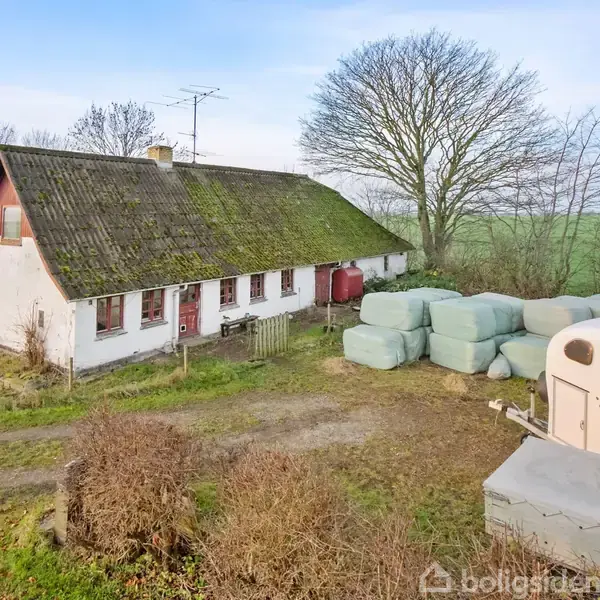 Et gammelt hus med mosdækket tag står omgivet af marker og træer. Ved siden af ligger stablede høballer og en hestevogn. Vinterbar jord dækker det meste af gårdspladsen.