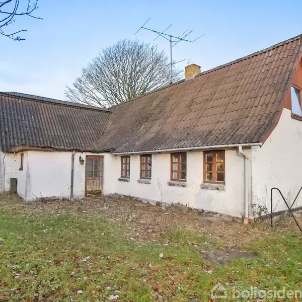 Et hvidt hus med brune skodder og skråt tag står i en have med græs og visne blade. Træer uden blade omgiver huset i baggrunden.