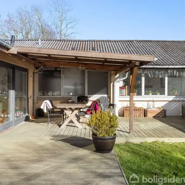 Terrassebord og stole står under en overdækket terrasse, omgivet af en græsplæne og trægulv. Store vinduer på huset giver udsigt til den solrige have.