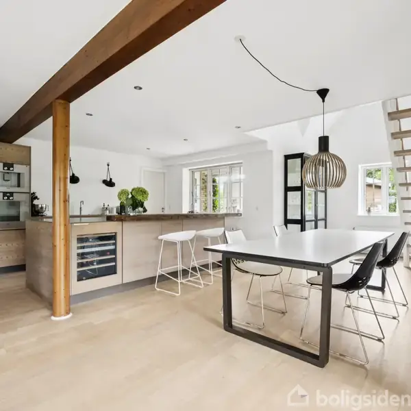 En moderne åben planløsning viser et køkken og spiseområde med et langt hvidt bord og minimalistiske stole. Trappe til højre, køkkenø til venstre, lyse trægulve.