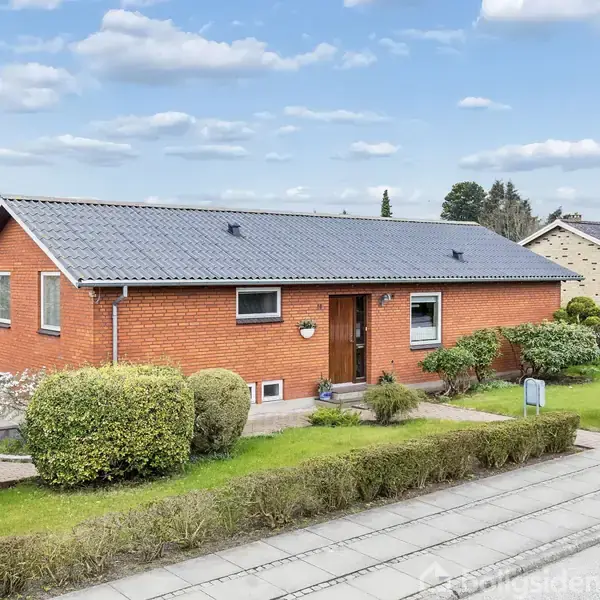 Rødt murstenshus med gråt saddeltag, omgivet af veltrimmet hæk og buske, placeret i en forstadslignende indstilling med flere huse i baggrunden under en blå skyet himmel.