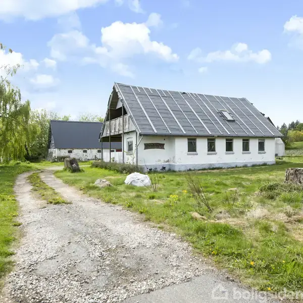 Et hvidt hus med skråtag og solpaneler står alene. Det ligger på en grusvej omgivet af græs og træer under en blå himmel med skyer.