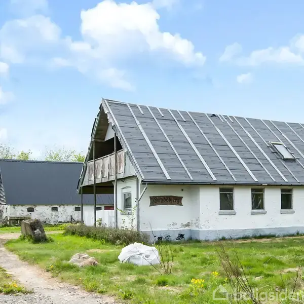 Hus med sort tag står stille på en grusvej omgivet af græs og træer, under en blå himmel. Anden bygning ses i baggrunden.