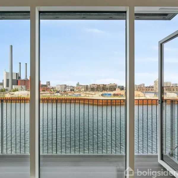 Åben altandør leder ud mod vandkanten. I baggrunden ses industri- og boligbygninger langs kysten under en blå himmel.