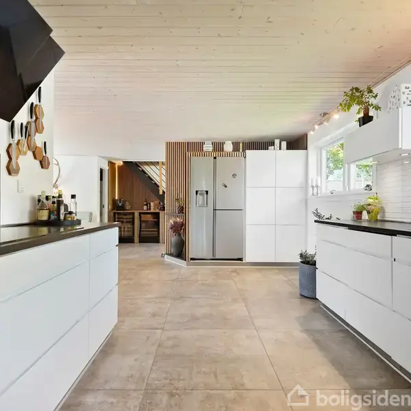 Et moderne køkken med hvide skabe og sorte bordplader står roligt. Der er en stål-køleskab i baggrunden ved siden af store vinduer, som slipper lys ind i rummet.