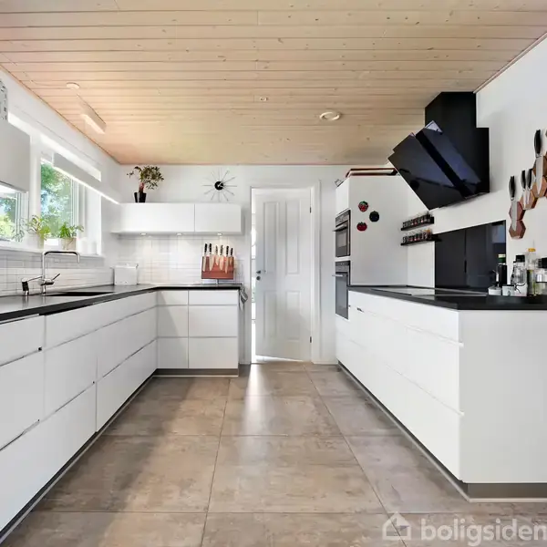 Et lyst, moderne køkken med hvide skabe og sorte bordplader. Køkkenet har en indbygget ovn, knivholder og dekoration på væggen. Store vinduer giver naturligt lys og udsigt til grønne områder.