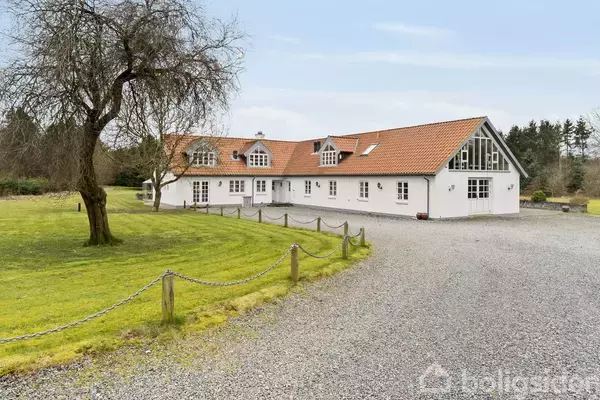 En hvid, langstrakt bygning med rødt tegltag står på en grusbelagt indkørsel, omgivet af en grøn græsplæne og enkelte træer under en blå himmel.