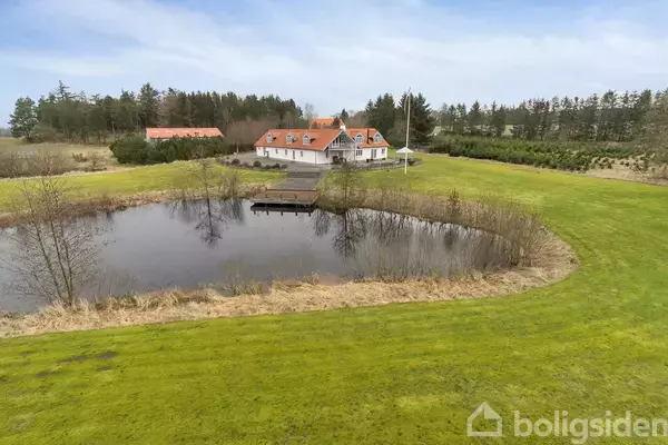 Hus med rødt tag og hvide vægge ligger ved siden af en sø med bro, omgivet af grønt græs og træer i baggrunden.