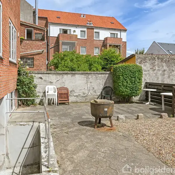 Stole og grill står stille på en flisebelagt terrasse omgivet af høje mure. Bagved ses en rød murstensbygning med rødt tag og flere vinduer, samt en hæk.