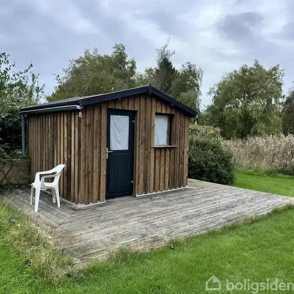 Et lille træhus står stille på en træterrasse. En hvid plastikstol er placeret ved siden af døren. Omgivet af grønt græs og høje buske.
