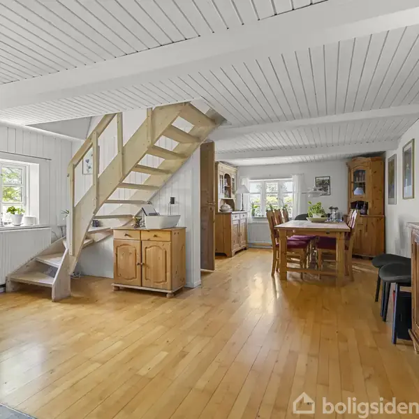 Trætrappe fører opad i en lys stue med træmøbler og spisebord til seks personer. Vinduerne lader naturligt lys strømme ind. Væggene og loftet er træpanelerede.