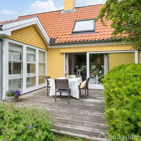 Et hus med gul væg og rødt tegltag har en terrasse med hvidt havesæt. Hækken og frodige planter omgiver området under en klar himmel.