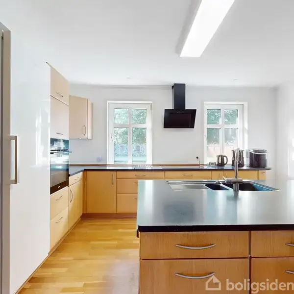 Et moderne køkken med sorte bordplader og lyse træskabe. Rummet er lyst med to vinduer og en række apparater placeret omkring vasken og kogepladen. En vase med blomster står på bordet.