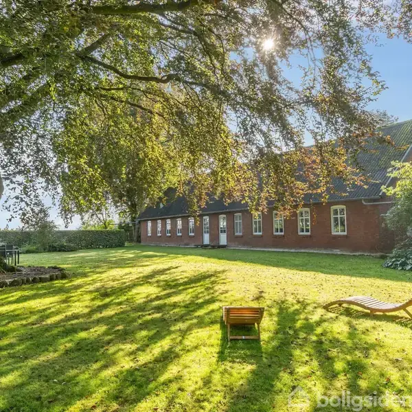 Plænestol står på solbeskinnet græsplæne foran stort træ. I baggrunden ses en lang, rød murstensbygning med mange vinduer. Sollys skaber skygger fra træerne.