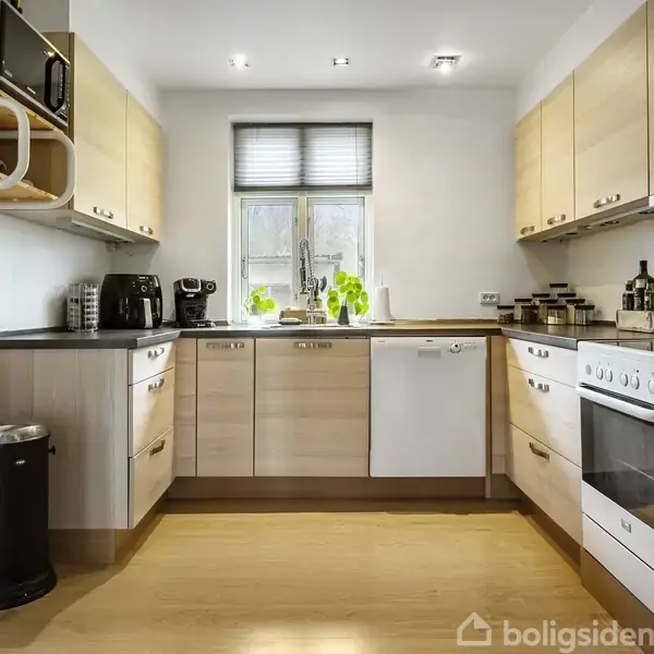 Et moderne køkken med lyse træskabe, to kaffemaskiner og en komfur. Planter står ved vinduet. Omgivet af en opvaskemaskine, mikrobølgeovn og organiserede krydderier. Lyst trægulv følger hele rummet.