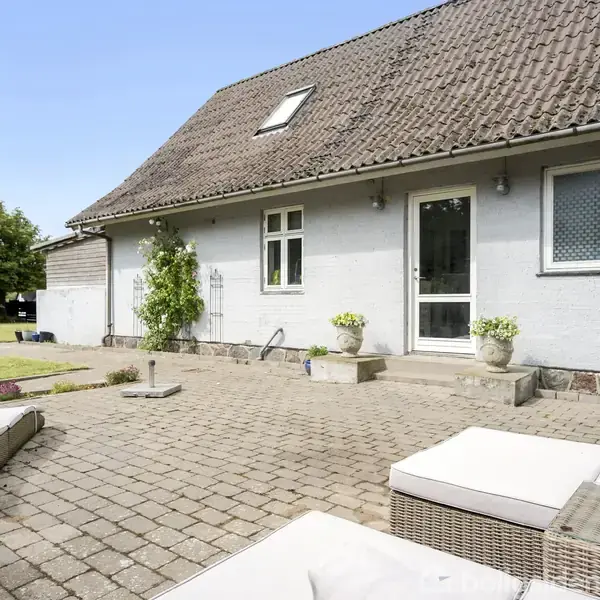 Huset har en stor terrasse med fliser og udendørs møbler. Det står i et grønt haveområde med buske og græsplæne. Huset har gråt tag og hvid malet facade.