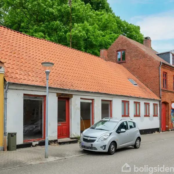 Gråt hus med røde vinduer og døre står stille ved en vej; en bil er parkeret foran; omgivet af træer og andre huse.
