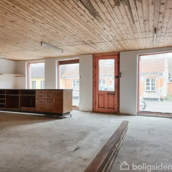 Et tomt rum med trægulv, indeholder en trækommode mod væggen. Indgangen er en rød dør med glas. Udenfor ses en facade af malede bygninger. Ingen tekst er til stede.