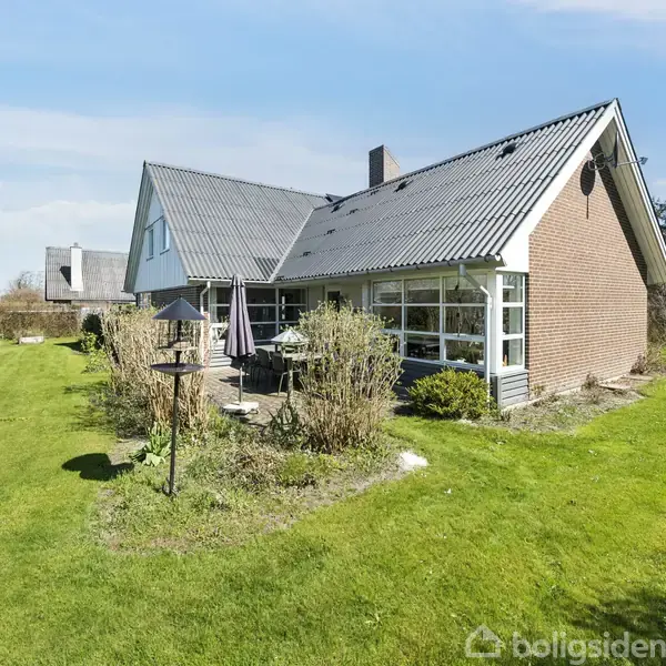 Et moderne, vinkelformet hus med røde murstensfacader og gråt tag står i en velplejet have med grønne græsplæner og buske. Et bord med parasol ses på terrassen.