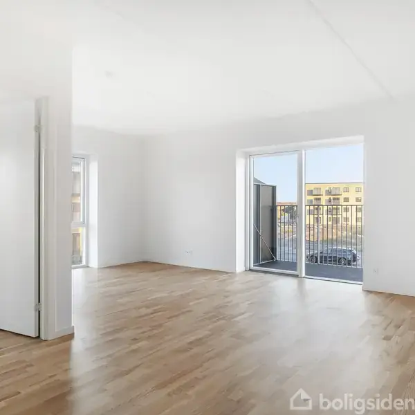 Et lyst, tomt rum med trægulv leder til en balkon gennem store skydedøre, omgivet af hvide vægge og skarpt lysindfald fra vinduerne i en moderne lejlighedskompleks.