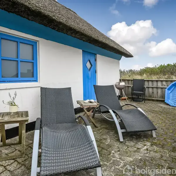 Et par sorte liggestole står på en flisebelagt terrasse ved siden af et hvidt hus med blå vinduesrammer og dør. Terrassen har et lille bord og en grill.