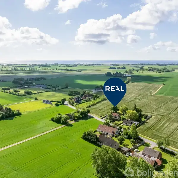 Luftfoto af landligt område: Marker i forskellige nuancer af grønt omkranser en samling huse. En blå markør med teksten "REA" peger på en bygning blandt dem.