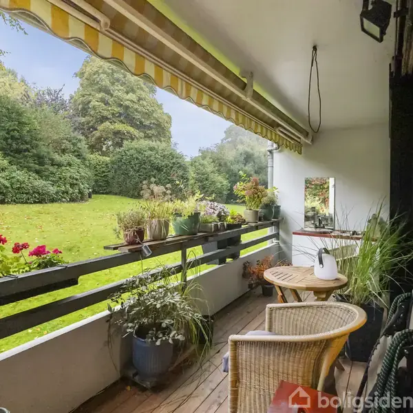 En terrasse med stole og planter, omgivet af grøn græsplæne og buske. Markisen er trukket ud, og der står en blomsterdekoration på altanrækværket.