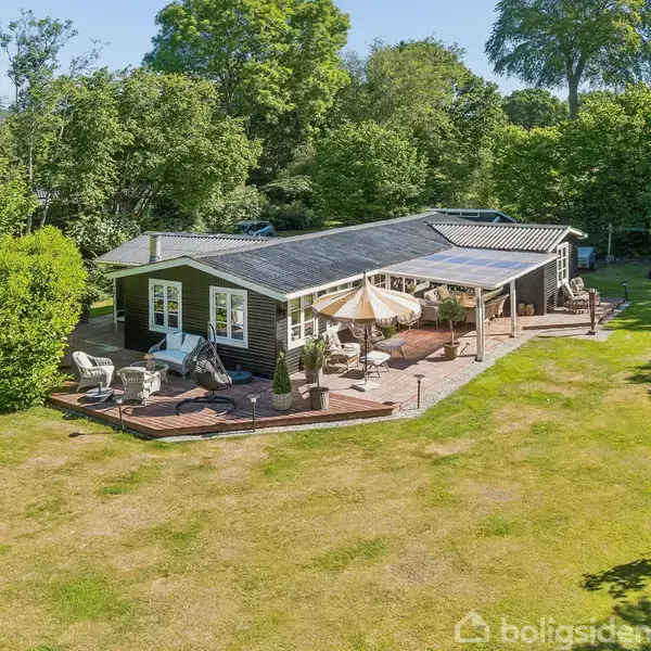 Et sommerhus står i en grøn have omgivet af træer. Terrassen har havemøbler og en stor parasol. Græsplænen breder sig foran huset, der er omgivet af frodigt løv.