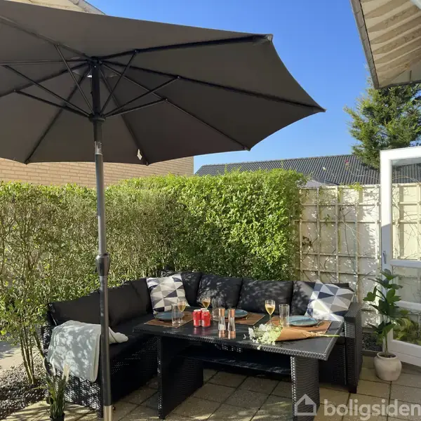 En sort parasol skygger over et udendørs opholdsområde med sofamøbler og et bord med drikkevarer og snacks. Omkranset af grønne hække og en gul murstensvæg under en klar blå himmel.