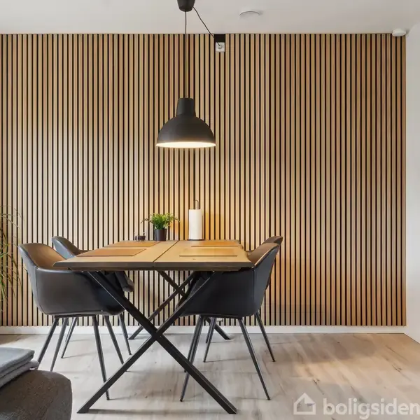 Et spisebord med sorte stole står i et moderne rum. En sort pendellampe hænger over bordet. Bagved er en væg med lodrette trælister. En grøn plante står ved siden af.