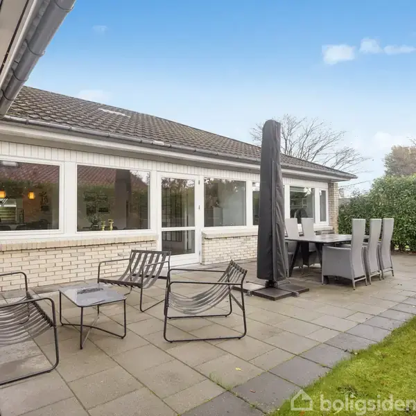 Terrasse med moderne møbler står stille foran et hus med store vinduer. Omkring terrassen er der en grøn græsplæne og buske. Himlen er let overskyet.