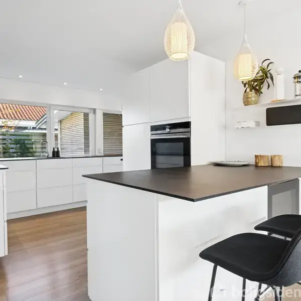 Et moderne køkken med hvidt interiør, hvor to sorte stole står ved en ø med sort bordplade. Overskabe indeholder planter og dekorative genstande. Store vinduer giver naturligt lys.