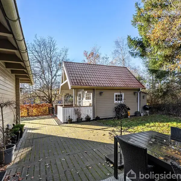 Et træhus står stille på en flisebelagt terrasse omgivet af træer og græsplæne. Ved siden af står et sort havebord med stole.