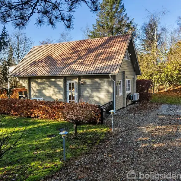 Et gråt hus med rødt tag står omgivet af træer og hække. Indkørslen er dækket af grus. Haven er grøn med enkelte lamper.