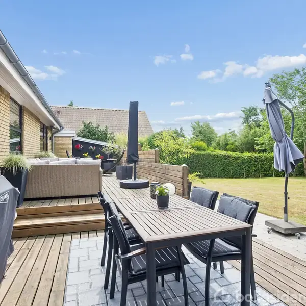 Terrasse med lang spisebord, sorte stole og planter. Grill står ved siden og en parasol er sammenklappet. I baggrunden ses en græsplæne og buske under en blå himmel.