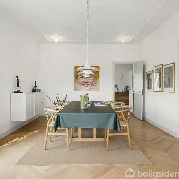Et spisebord med seks stole står centreret i et lyst rum med trægulv. Et stort maleri hænger på væggen, og der er en lampe over bordet.