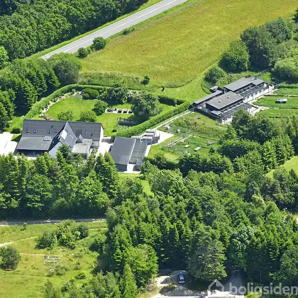 Et stort hus med sorte tage omgivet af træer står i et grønt landskab ved en landevej. Flere bygninger og velplejede haver ses omkring hovedstrukturen.