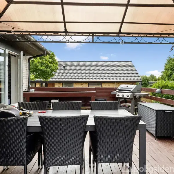 Terrassemøblement består af et bord med seks sorte stole under et halvtag. En grill står ved siden af, og omgivelserne inkluderer grønne træer og nabohuse.
